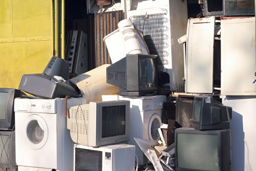 A collection of discarded household appliances and electronic waste arranged outdoors, including washing machines, microwaves, desktop computer monitors, and small fridge units, stacked closely together against a metal background. The appliances display various finishes and materials, such as white painted metal and plastic, with some appearing weathered or aged. In the foreground, a washing machine with a round door is positioned near a microwave oven and a computer monitor, both showing reflective screens and plastic casings. The overall scene suggests a large-scale rubbish collection effort, typical of commercial waste removal services operating in the Knightsbridge area. The environment appears to be an outdoor storage or collection site, with natural light illuminating the cluttered assortment of electrical waste, indicative of efforts by Commercial Waste Knightsbridge to clear bulky household rubbish efficiently.